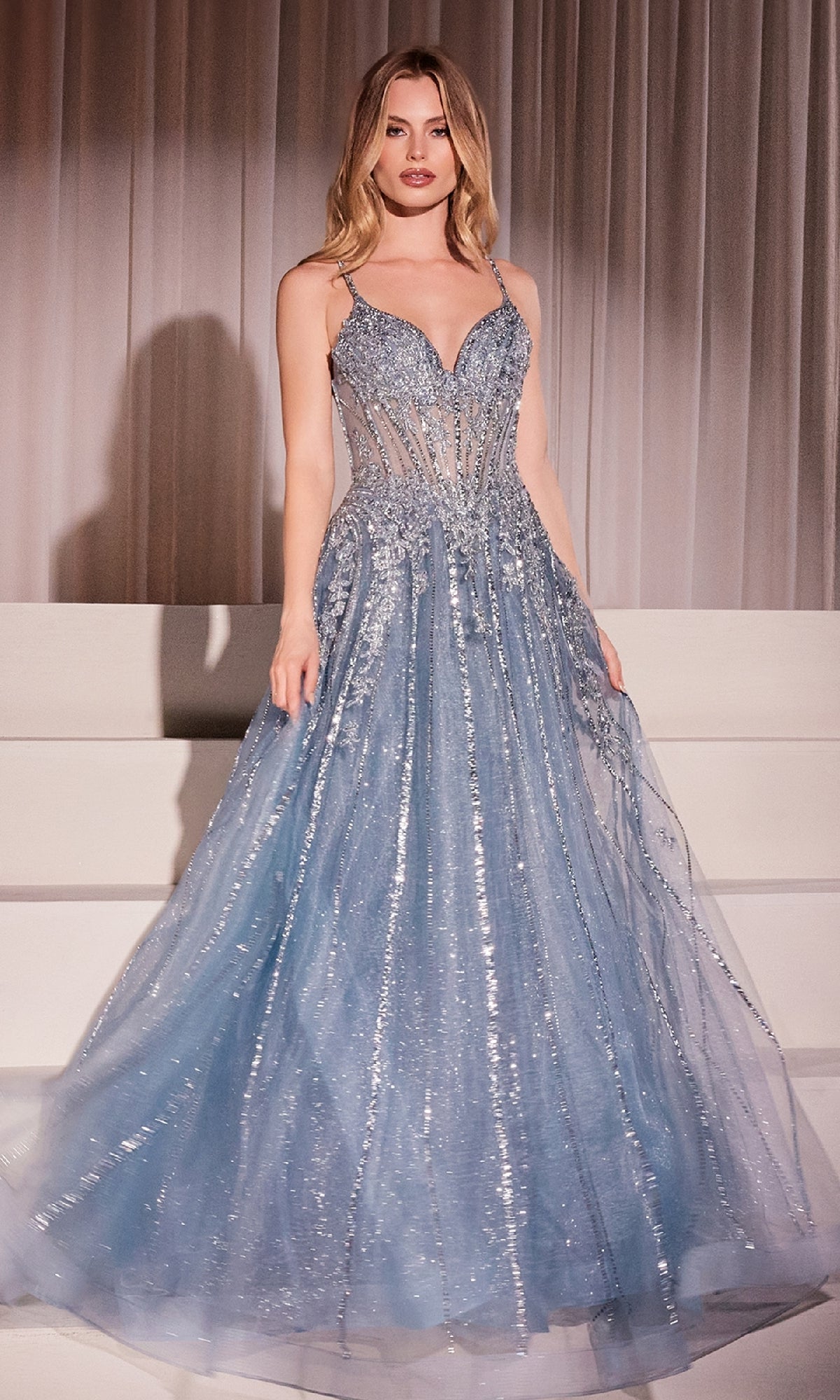 A woman stands on white steps in front of white curtains, wearing the Ladivine CD786 Crystal-Beaded Long Prom Dress—a light blue, floor-length gown with a fitted bodice and sheer panels.